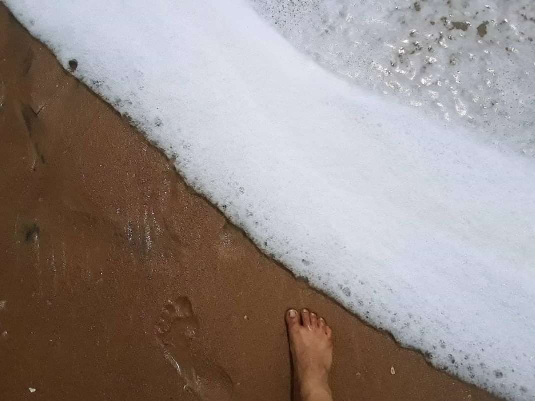 A woman's right foot and the impression of the left foot side by side on wet brown sand as seawater contaminated with industrial foam approaches from the top right corner